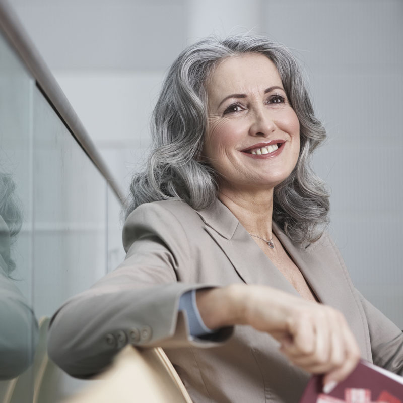 Middle-aged caucasian woman with long silver-gray hair, dressed in business attire and smiling, sitting in a chair with her arm draped on the back of the chair.