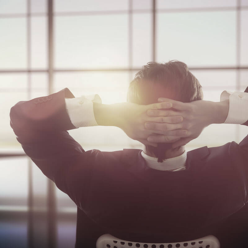 Back of a man dressed in a suit sitting in a chair leaning back slightly with his hands clasped on the back of his head. He is facing large windows with some sunlight peeking through towards him.