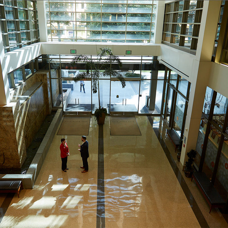 Wide shot looking down onto two adults standing and talking to each other in the large lobby of the interior of a building as the sun shies through the front windows.