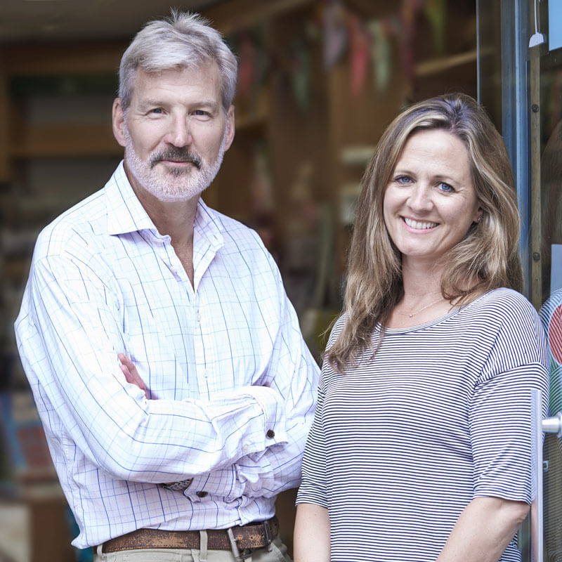 Two caucasian middle-aged adults smiling in the doorway of a business, a man with gray hair, beard, and mustache on the left, and a woman with long brown and gray hair on the right.